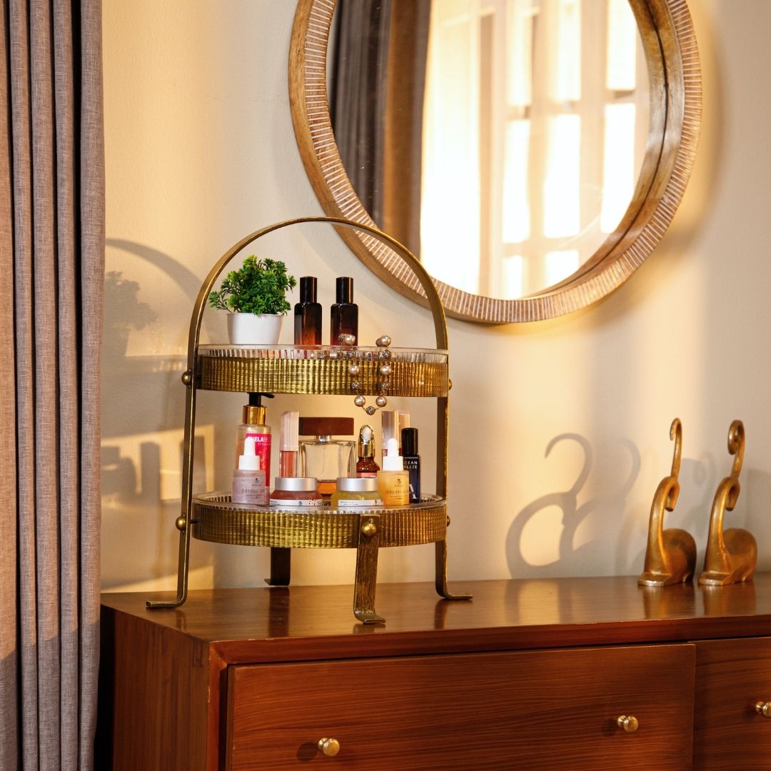 Two-tiered gold metal organizer placed on a wooden dresser beneath a round wall mirror, holding skincare and beauty products, with decorative golden figurines and a photo frame on the side.