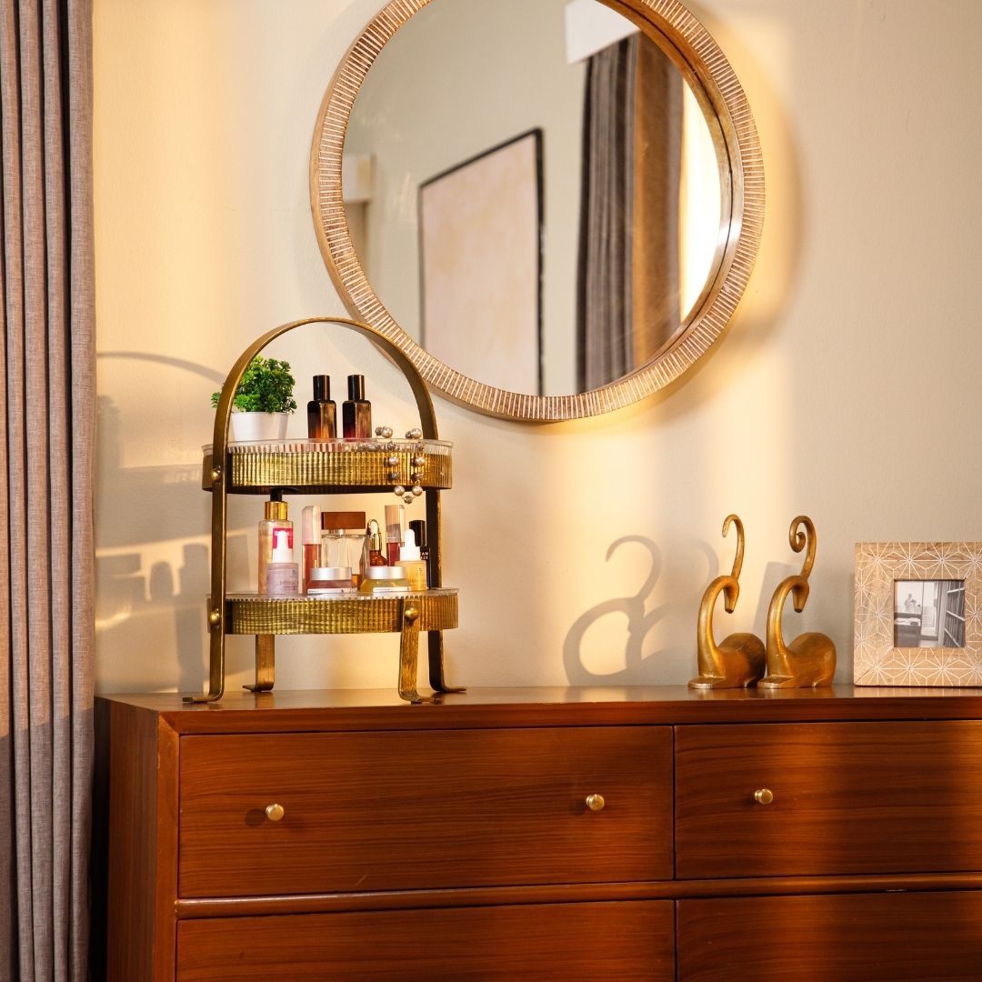 Two-tiered gold metal organizer placed on a wooden dresser beneath a round wall mirror, holding skincare and beauty products, with decorative golden figurines and a photo frame on the side.
