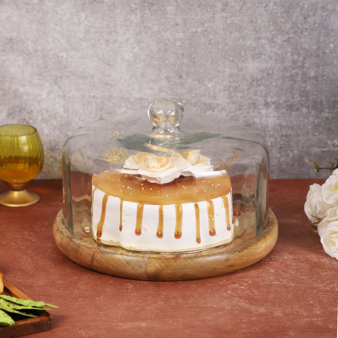 Hand lifting a clear glass dome off a beautifully decorated cake placed on a round wooden base, surrounded by snacks and flowers