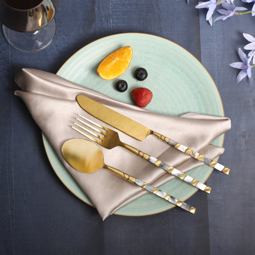 A decorative table setting featuring gold flatware with silver handles on a beige napkin, placed on a light blue ceramic plate, accompanied by fruit arranged to resemble a face with blueberries, a strawberry, and an orange slice