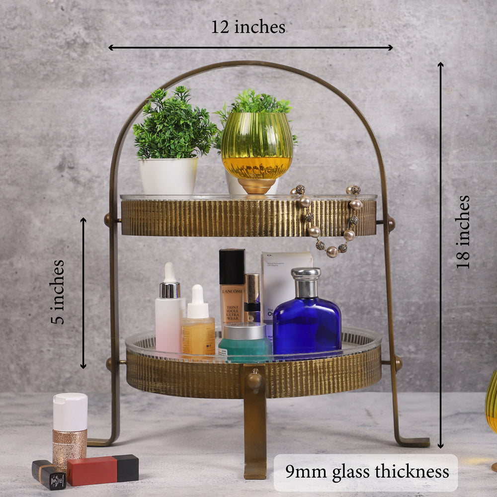 Two-tiered vintage-style metal organizer with ribbed gold finish, holding skincare bottles, a blue perfume bottle, a yellow candleholder, and small potted plants, placed on a marble surface.