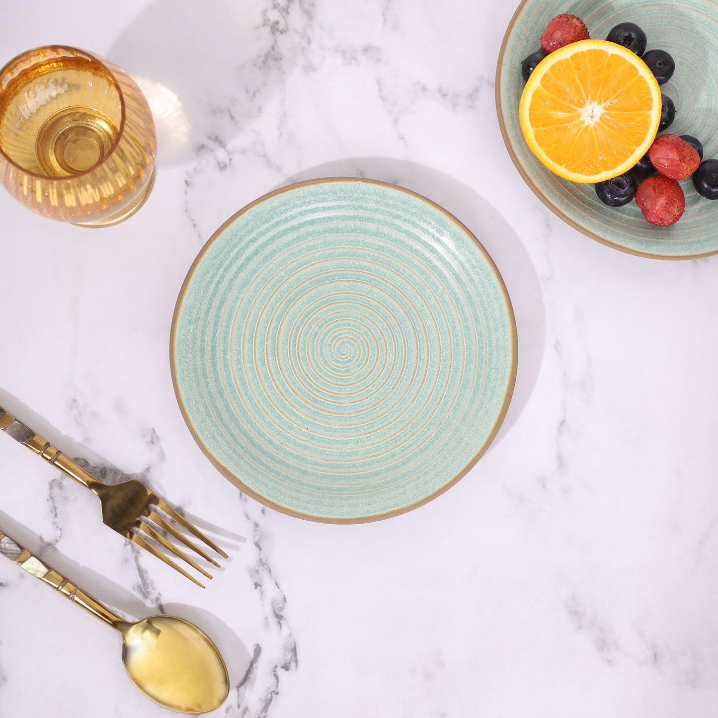 Green ceramic plate with a spiral design, placed on a marble surface alongside a gold spoon, fork, and a glass