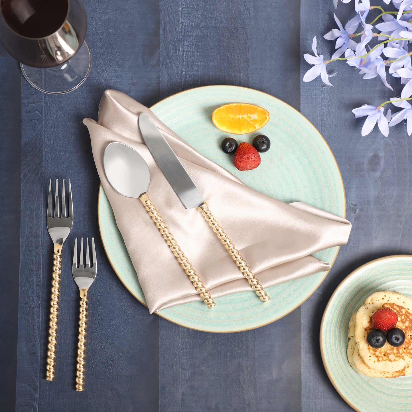 A decorative table setting featuring gold flatware with silver handles on a beige napkin, placed on a light blue ceramic plate, accompanied by fruit arranged to resemble a face with blueberries, a strawberry, and an orange slice