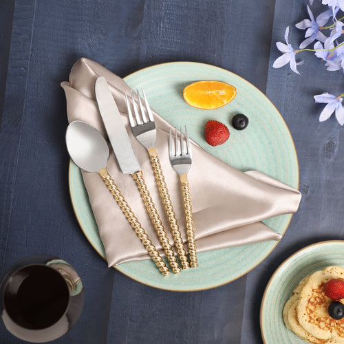 A decorative table setting featuring gold flatware with silver handles on a beige napkin, placed on a light blue ceramic plate, accompanied by fruit arranged to resemble a face with blueberries, a strawberry, and an orange slice
