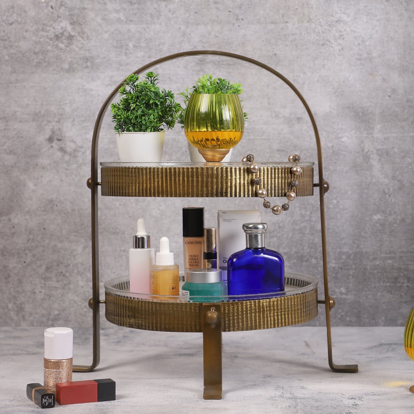 Two-tiered vintage-style metal organizer with ribbed gold finish, holding skincare bottles, a blue perfume bottle, a yellow candleholder, and small potted plants, placed on a marble surface.