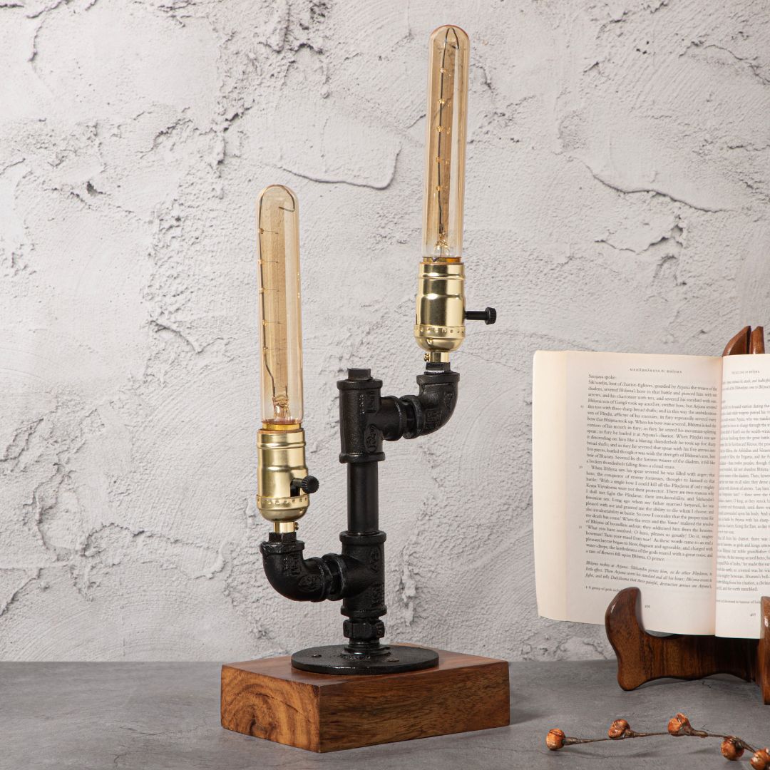 Industrial-style table lamp with a pipe-inspired design, dual brass-tone bulb holders, and a wooden base, displayed alongside greenery and an open book on a concrete-textured background