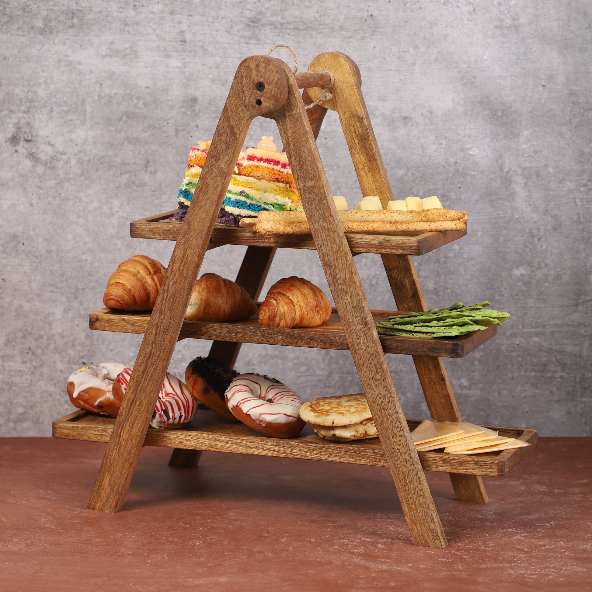 Three-tier wooden serving stand with an A-frame design, displaying assorted pastries, rainbow cake slices, crackers, and leafy greens on a brown tabletop with a neutral gray background
