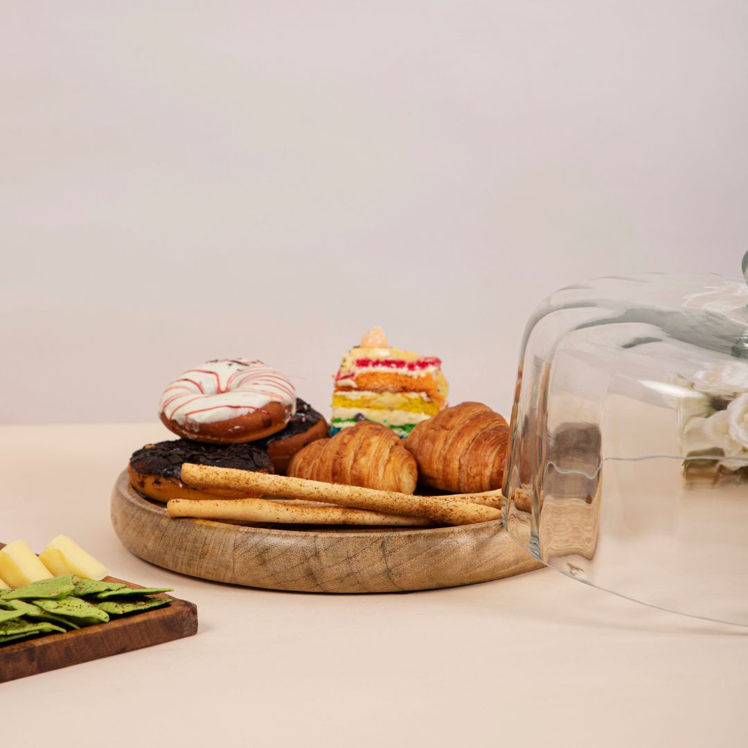 A variety of pastries and desserts, including donuts, croissants, and cake, displayed on a wooden platter with a glass cloche