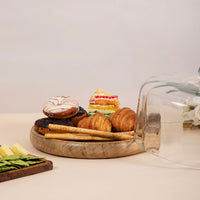 A variety of pastries and desserts, including donuts, croissants, and cake, displayed on a wooden platter with a glass cloche — thumbnail