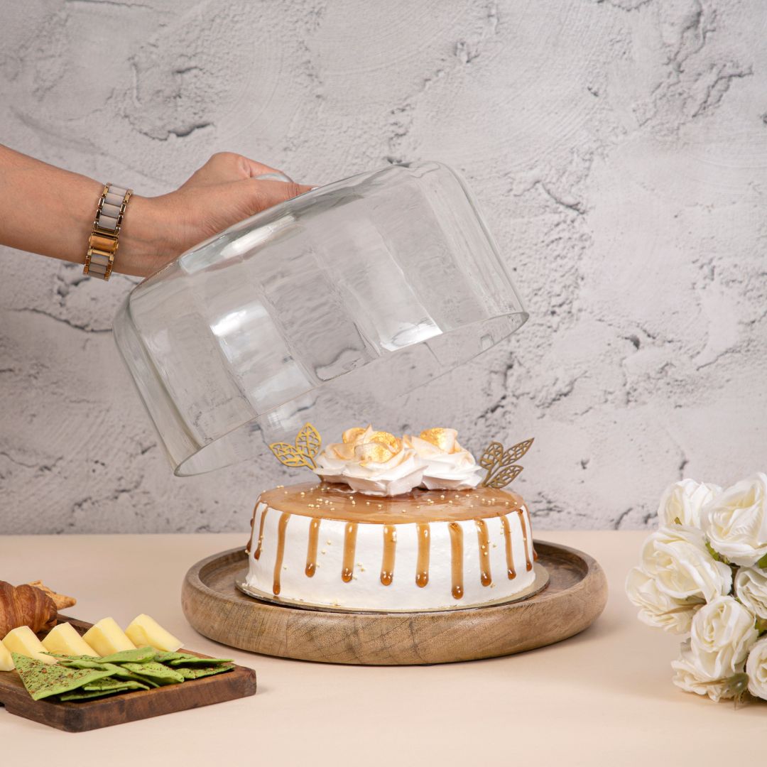 Hand lifting a clear glass dome off a beautifully decorated cake placed on a round wooden base, surrounded by snacks and flowers