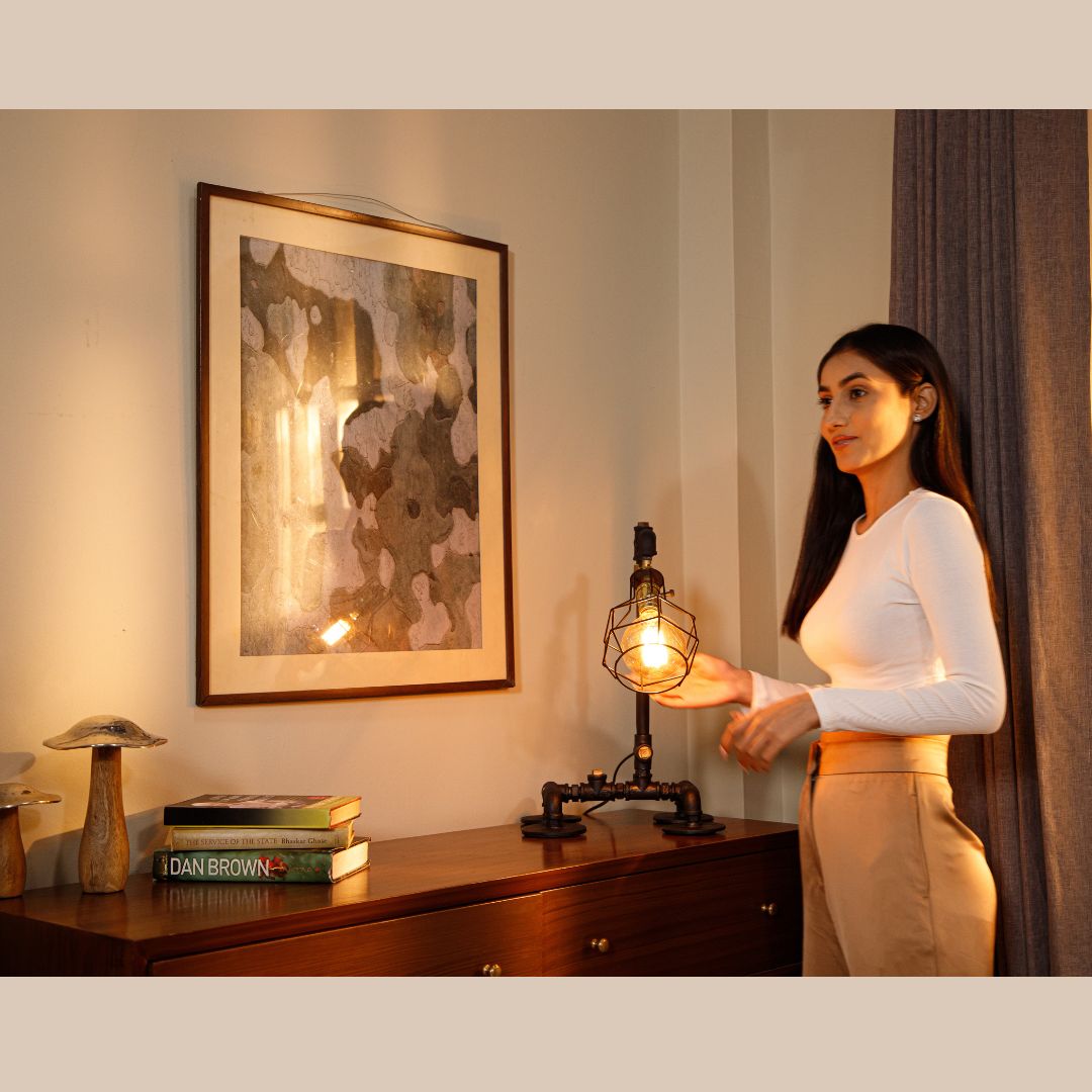 A woman in a well-lit room standing beside an industrial-style lamp on a wooden table