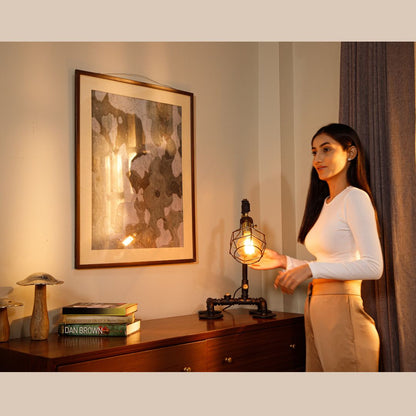 A woman in a well-lit room standing beside an industrial-style lamp on a wooden table