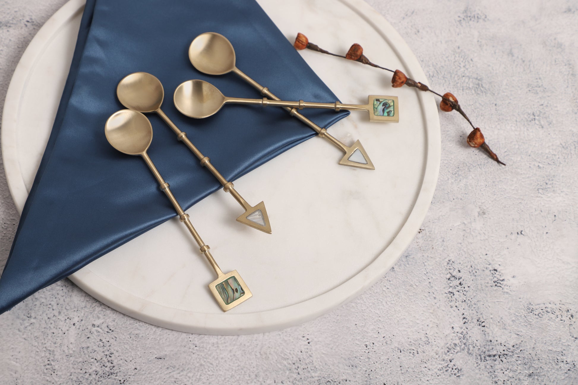  Elegant gold spoons with decorative geometric handles featuring abalone inlays, placed on a folded navy blue napkin atop a round white marble tray, accompanied by a branch with small brown pods