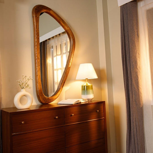 Wooden-framed asymmetrical mirror above a wooden dresser, styled with a white vase and a lamp