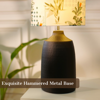 Table lamp with a dark, textured metal base and a floral-patterned fabric shade, placed on a white table in a room with plants — thumbnail