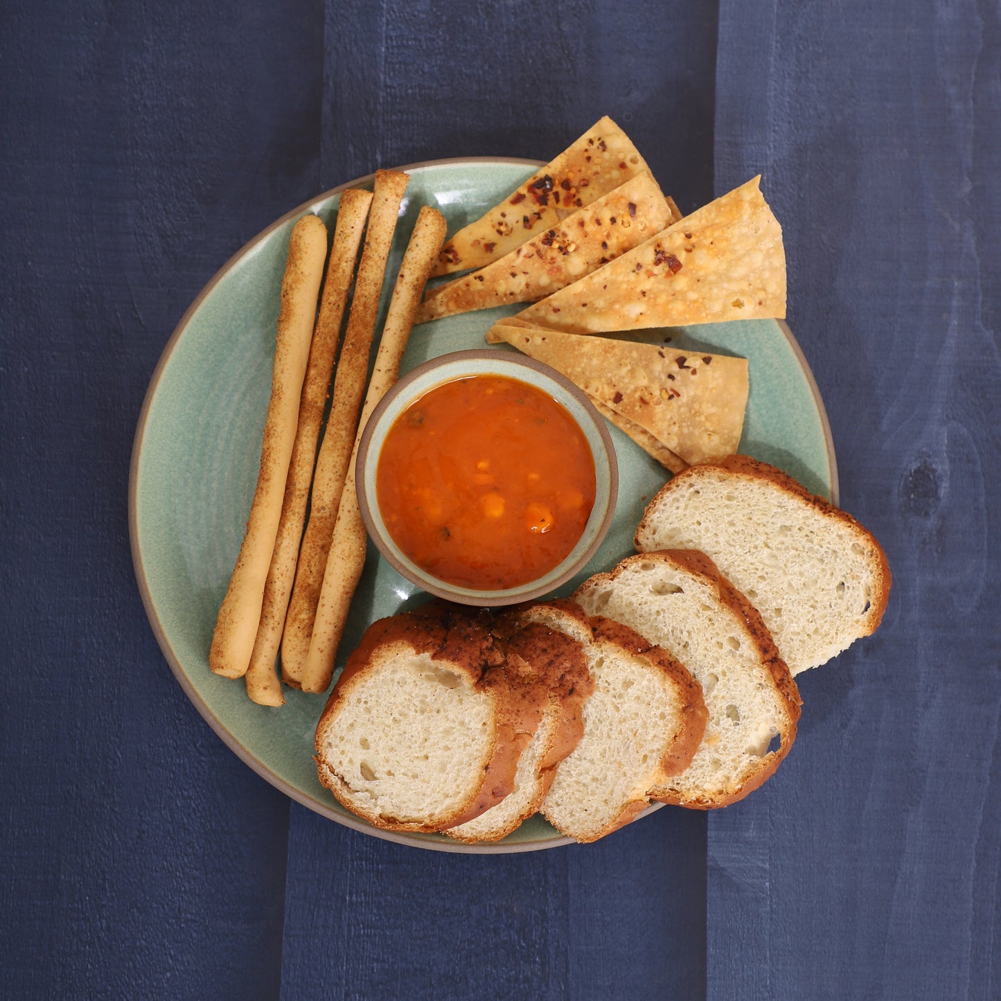 Chips-and-dip platter with breadsticks, skewers, tortilla chips, cucumber slices, and red sauce, served alongside a croissant and pancakes on a marble and earthy background