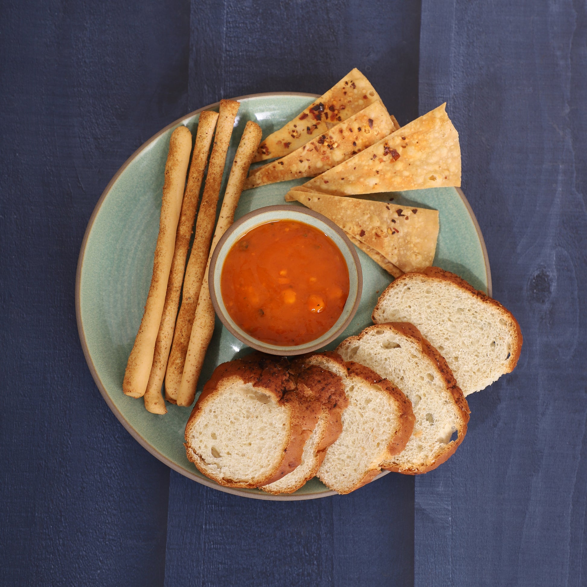 Chips-and-dip platter with breadsticks, skewers, tortilla chips, cucumber slices, and red sauce, served alongside a croissant and pancakes on a marble and earthy background