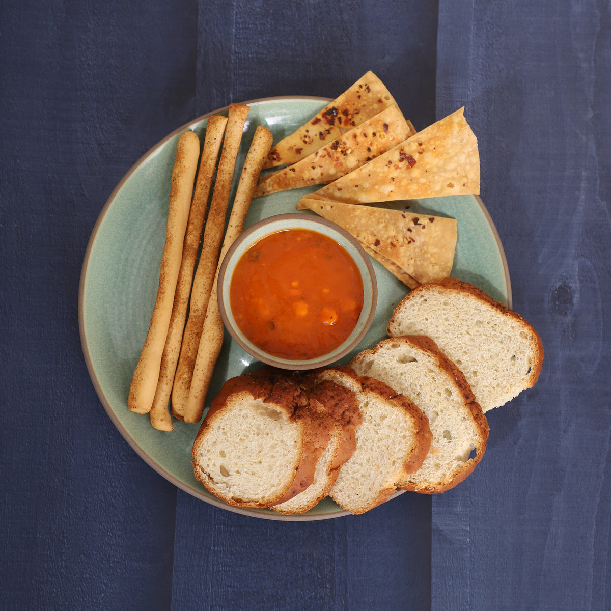 Chips-and-dip platter with breadsticks, skewers, tortilla chips, cucumber slices, and red sauce, served alongside a croissant and pancakes on a marble and earthy background