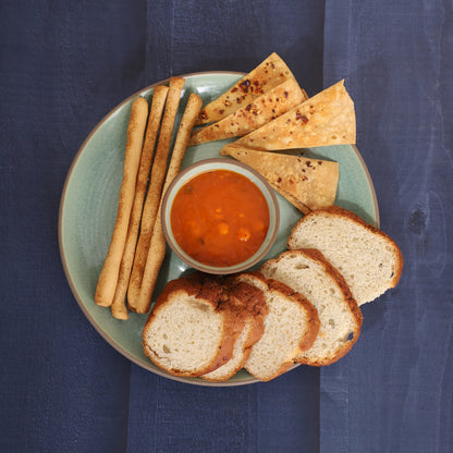 Chips-and-dip platter with breadsticks, skewers, tortilla chips, cucumber slices, and red sauce, served alongside a croissant and pancakes on a marble and earthy background