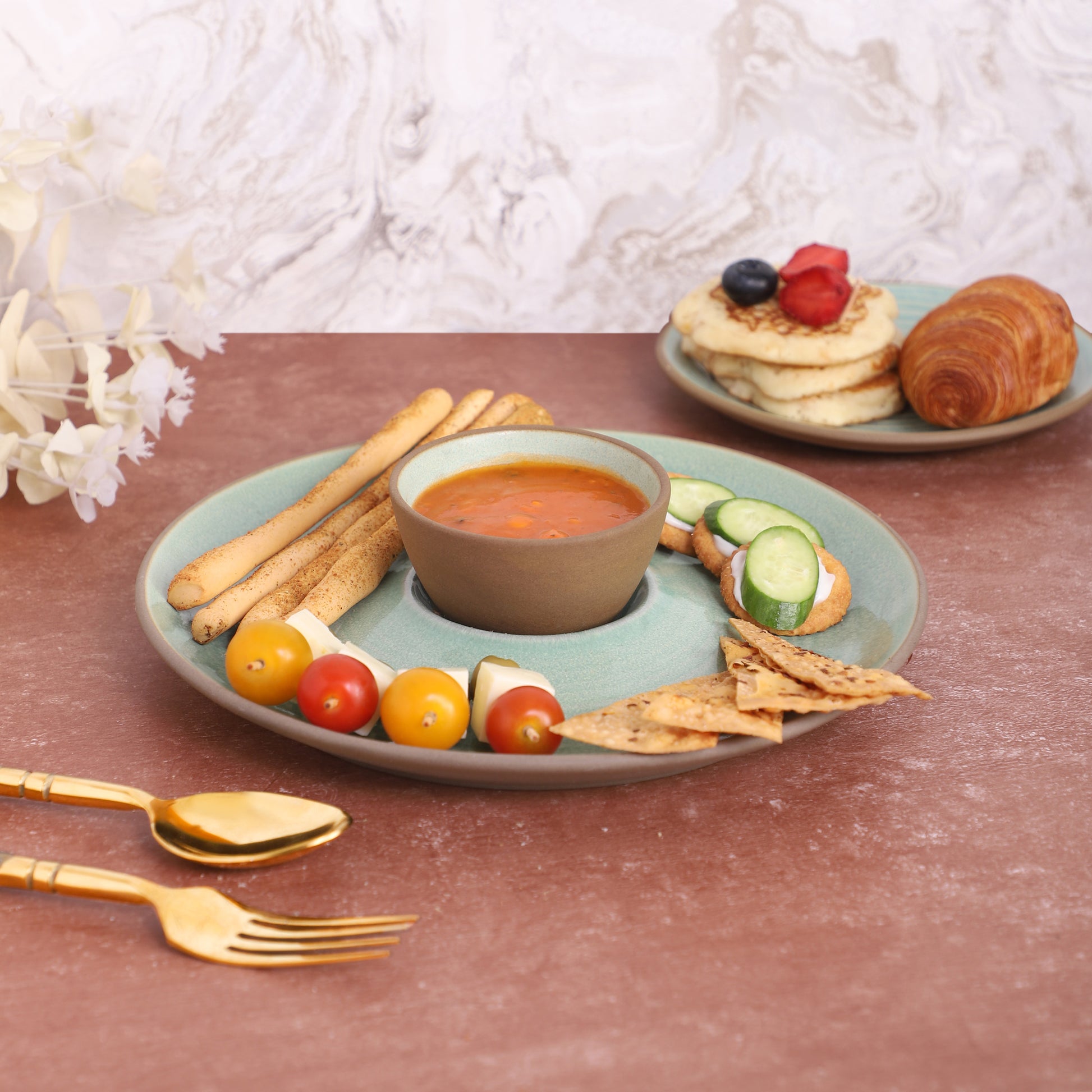 Chips-and-dip platter with breadsticks, skewers, tortilla chips, cucumber slices, and red sauce, served alongside a croissant and pancakes on a marble and earthy background