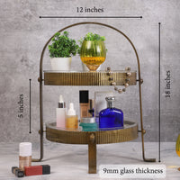 Two-tiered vintage-style metal organizer with ribbed gold finish, holding skincare bottles, a blue perfume bottle, a yellow candleholder, and small potted plants, placed on a marble surface. — thumbnail