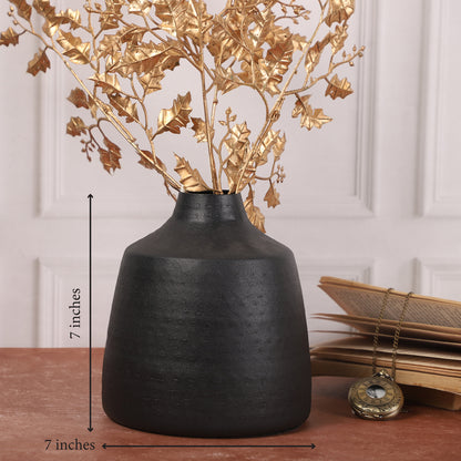 Black ceramic vase with golden eucalyptus stems placed on a red-brown table, accompanied by a small wooden jewelry box and stacked books in the background