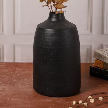 Black ceramic vase with golden eucalyptus stems placed on a red-brown table, accompanied by a small wooden jewelry box and stacked books in the background