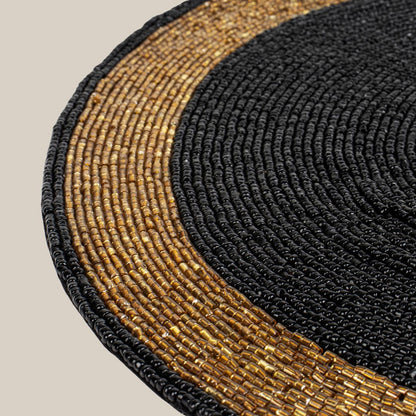 Round handcrafted beaded placemat with a black center and golden border, displayed on a concrete surface alongside
