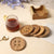 Wooden coasters with button-inspired designs and a matching holder, paired with a textured glass mug of tea and a golden spoon on a neutral tabletop