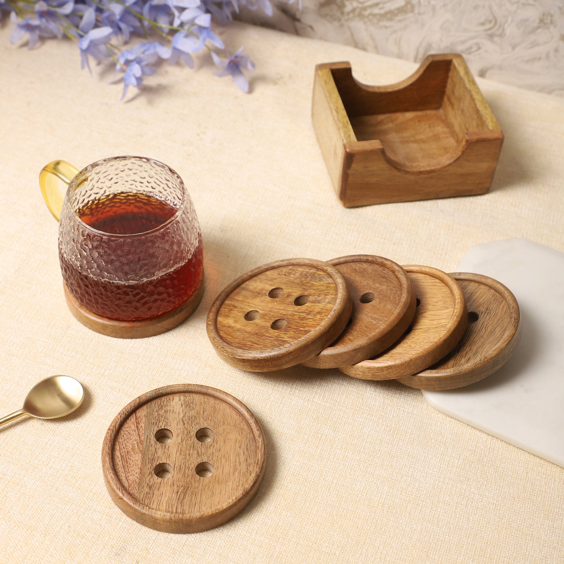 Wooden coasters with button-inspired designs and a matching holder, paired with a textured glass mug of tea and a golden spoon on a neutral tabletop