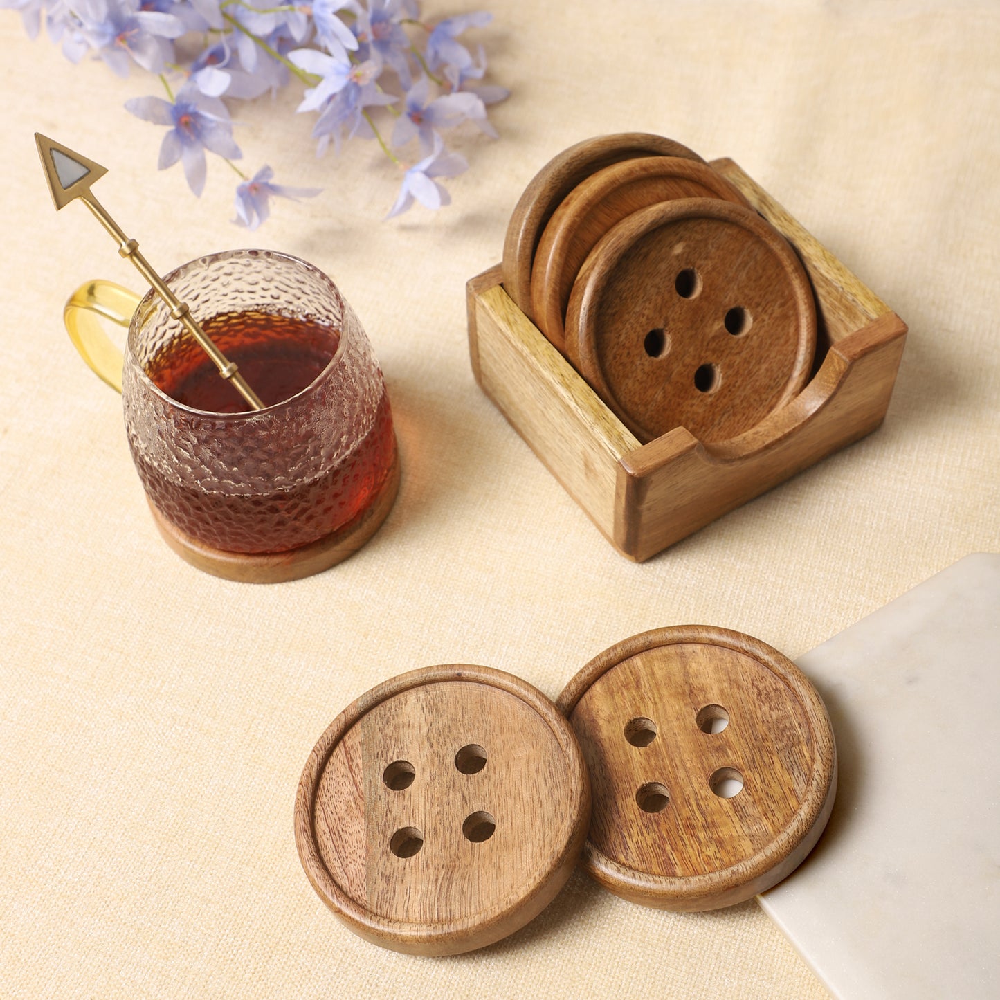 Wooden coasters with button-inspired designs and a matching holder, paired with a textured glass mug of tea and a golden spoon on a neutral tabletop
