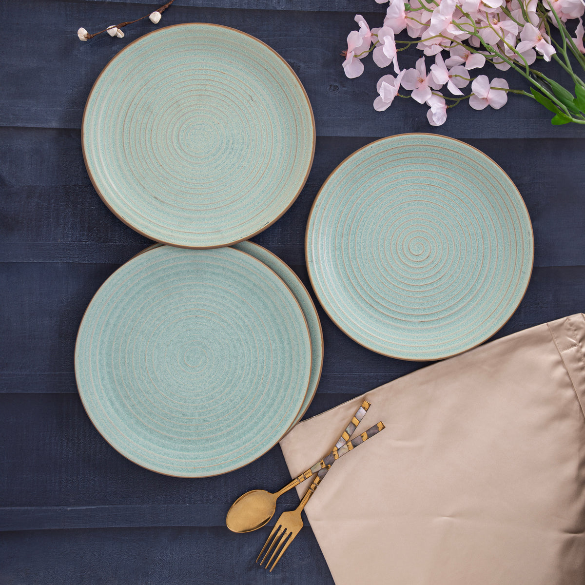 Three turquoise textured dinner plates arranged on a dark wooden surface with a beige napkin, gold cutlery, and pink flowers