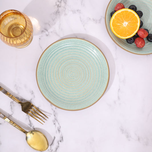 Green ceramic plate with a spiral design, placed on a marble surface alongside a gold spoon, fork, and a glass