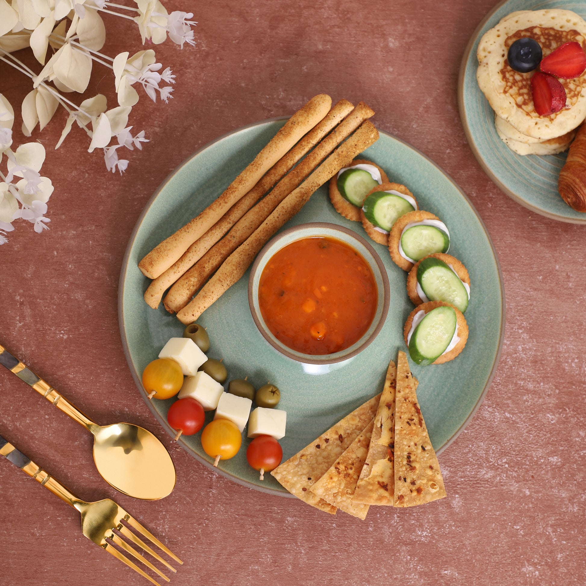 Chips-and-dip platter with breadsticks, skewers, tortilla chips, cucumber slices, and red sauce, served alongside a croissant and pancakes on a marble and earthy background