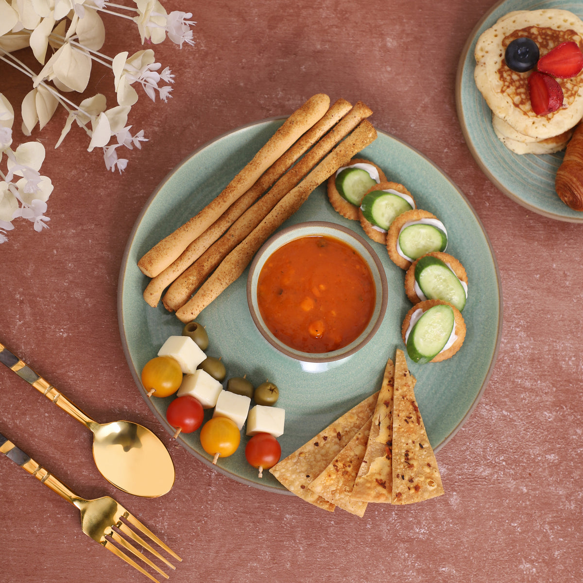 Chips-and-dip platter with breadsticks, skewers, tortilla chips, cucumber slices, and red sauce, served alongside a croissant and pancakes on a marble and earthy background