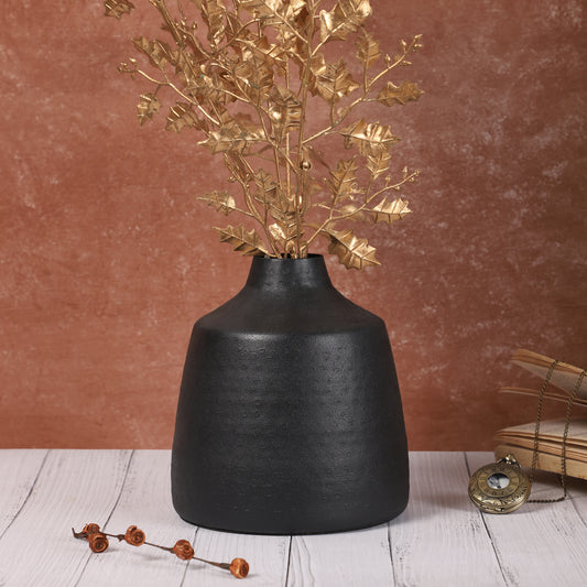Black ceramic vase with golden eucalyptus stems placed on a red-brown table, accompanied by a small wooden jewelry box and stacked books in the background