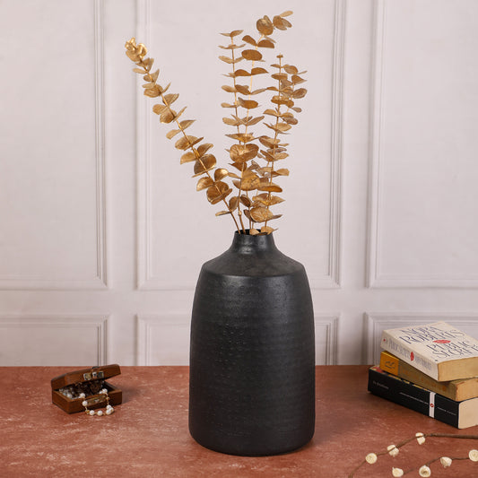 Black ceramic vase with golden eucalyptus stems placed on a red-brown table, accompanied by a small wooden jewelry box and stacked books in the background