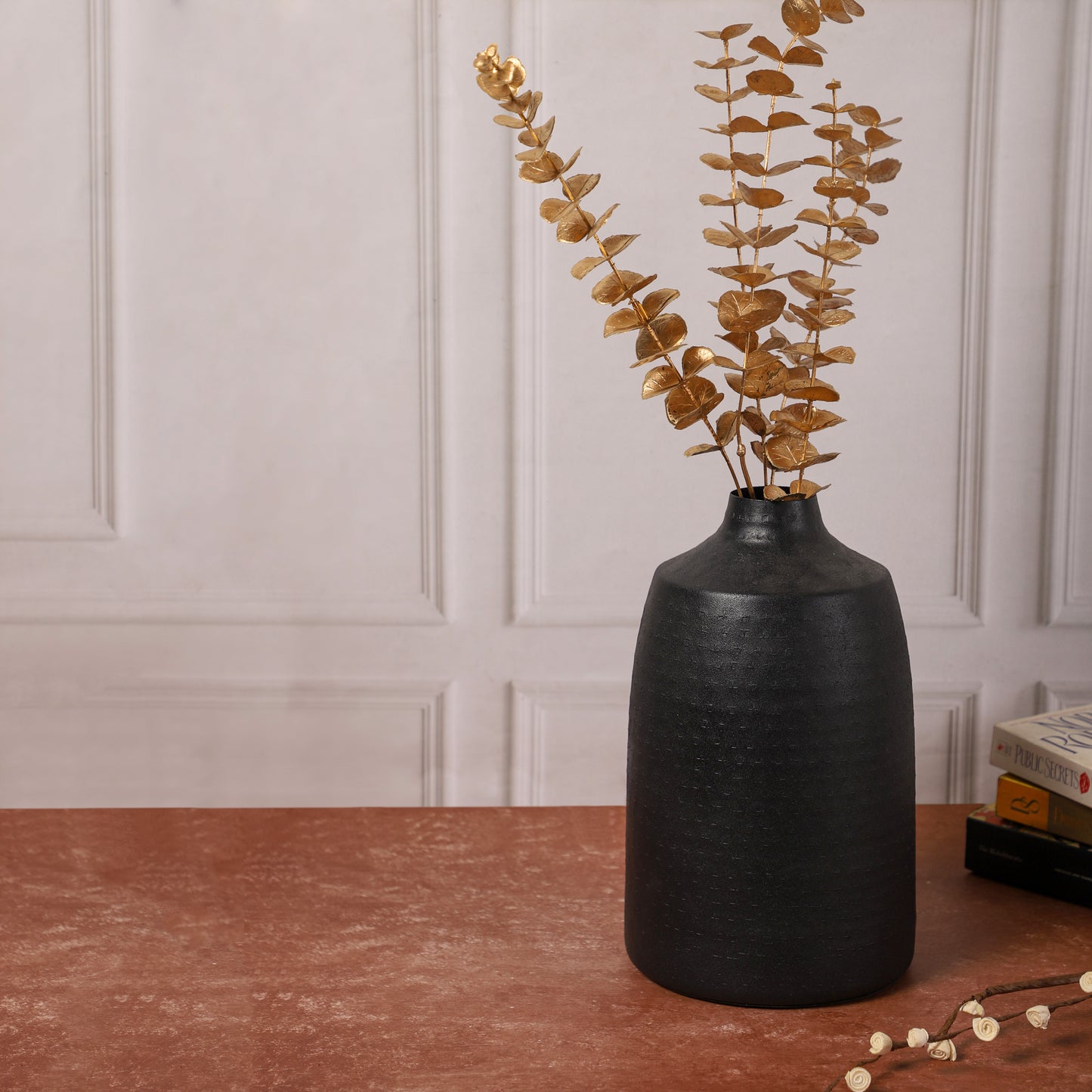 Black ceramic vase with golden eucalyptus stems placed on a red-brown table, accompanied by a small wooden jewelry box and stacked books in the background