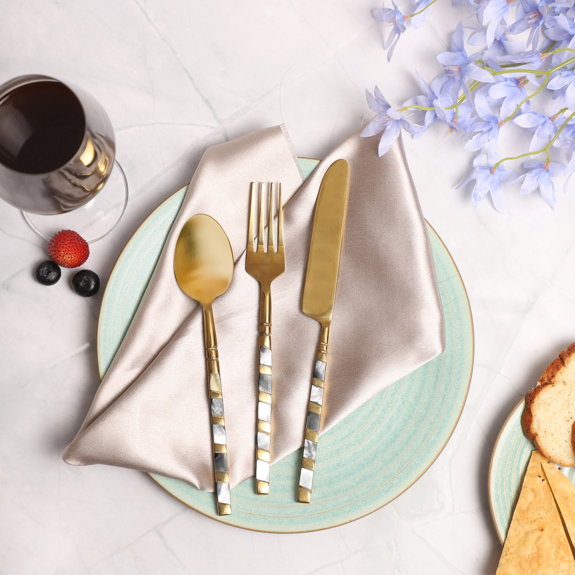 A decorative table setting featuring gold flatware with silver handles on a beige napkin, placed on a light blue ceramic plate, accompanied by fruit arranged to resemble a face with blueberries, a strawberry, and an orange slice