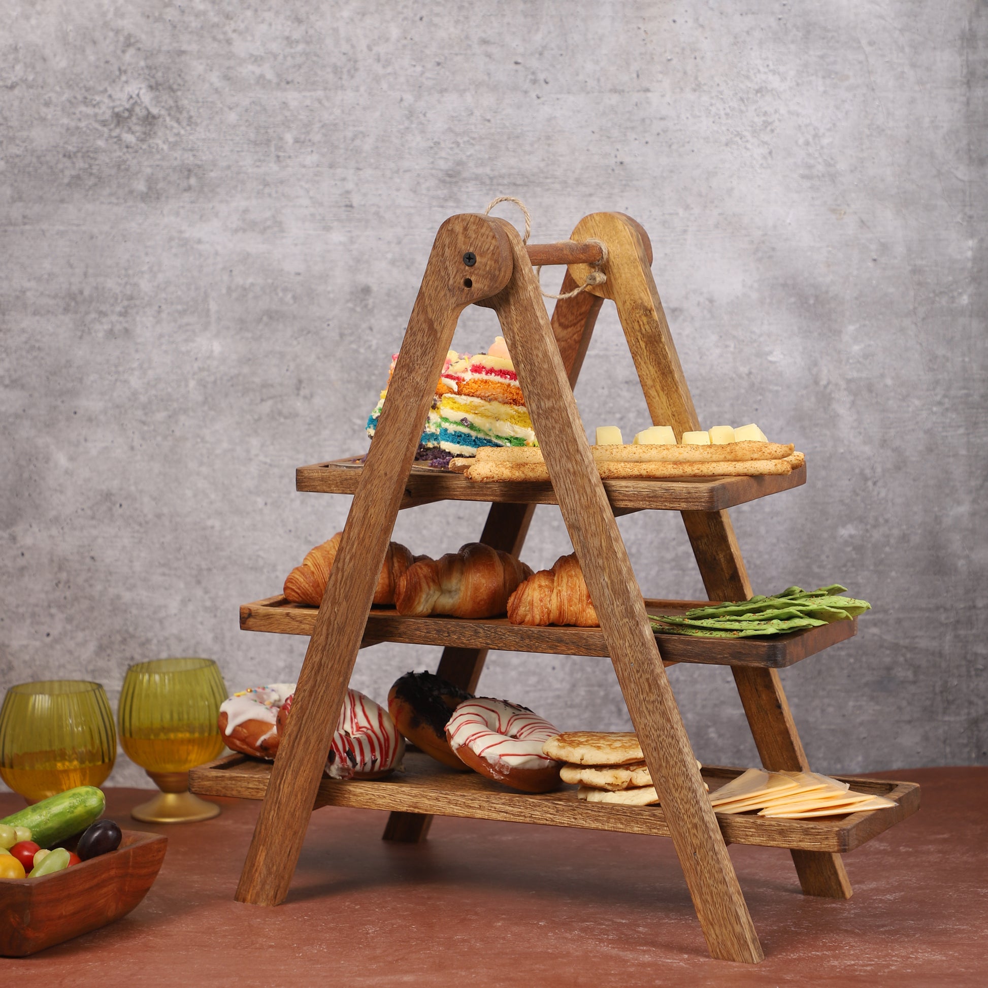Three-tier wooden serving stand with an A-frame design, displaying assorted pastries, rainbow cake slices, crackers, and leafy greens on a brown tabletop with a neutral gray background