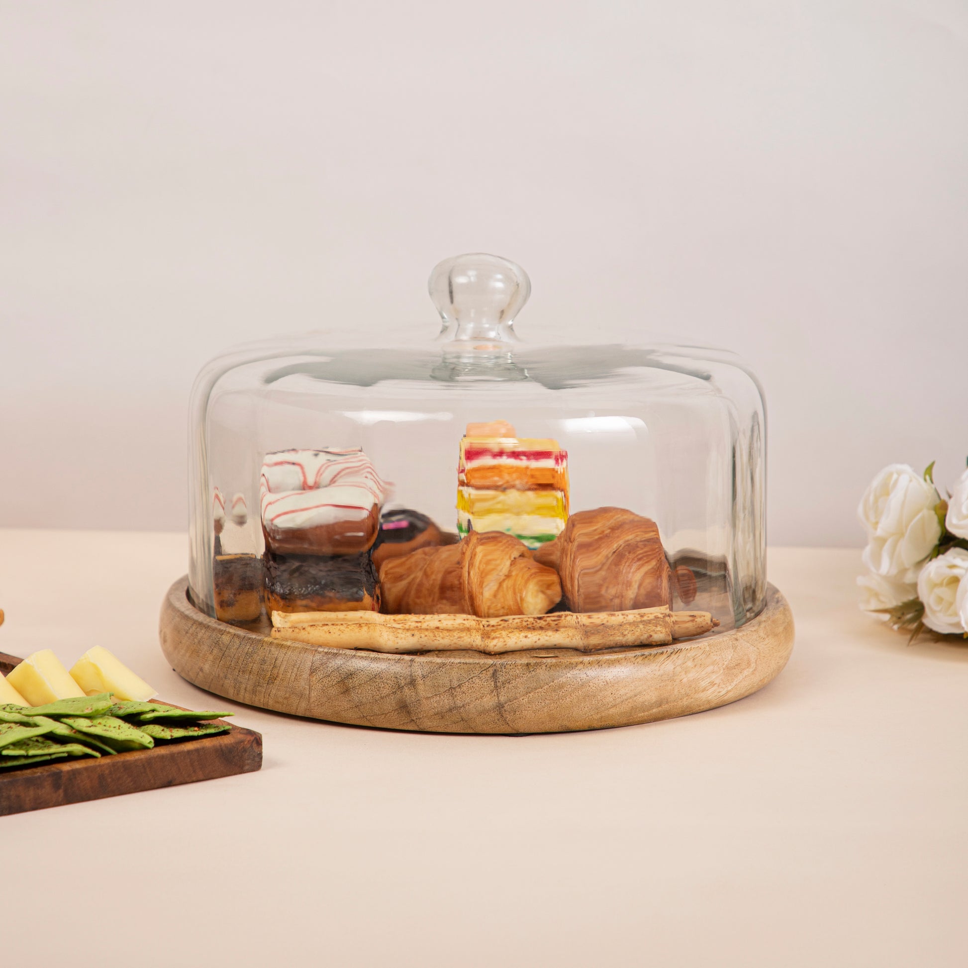 A variety of pastries and desserts, including donuts, croissants, and cake, displayed on a wooden platter with a glass cloche