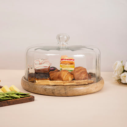 A variety of pastries and desserts, including donuts, croissants, and cake, displayed on a wooden platter with a glass cloche