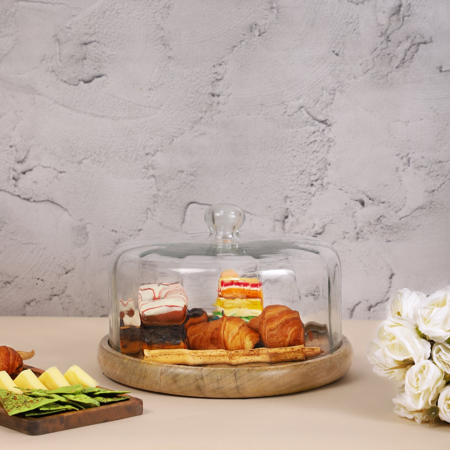 A variety of pastries and desserts, including donuts, croissants, and cake, displayed on a wooden platter with a glass cloche
