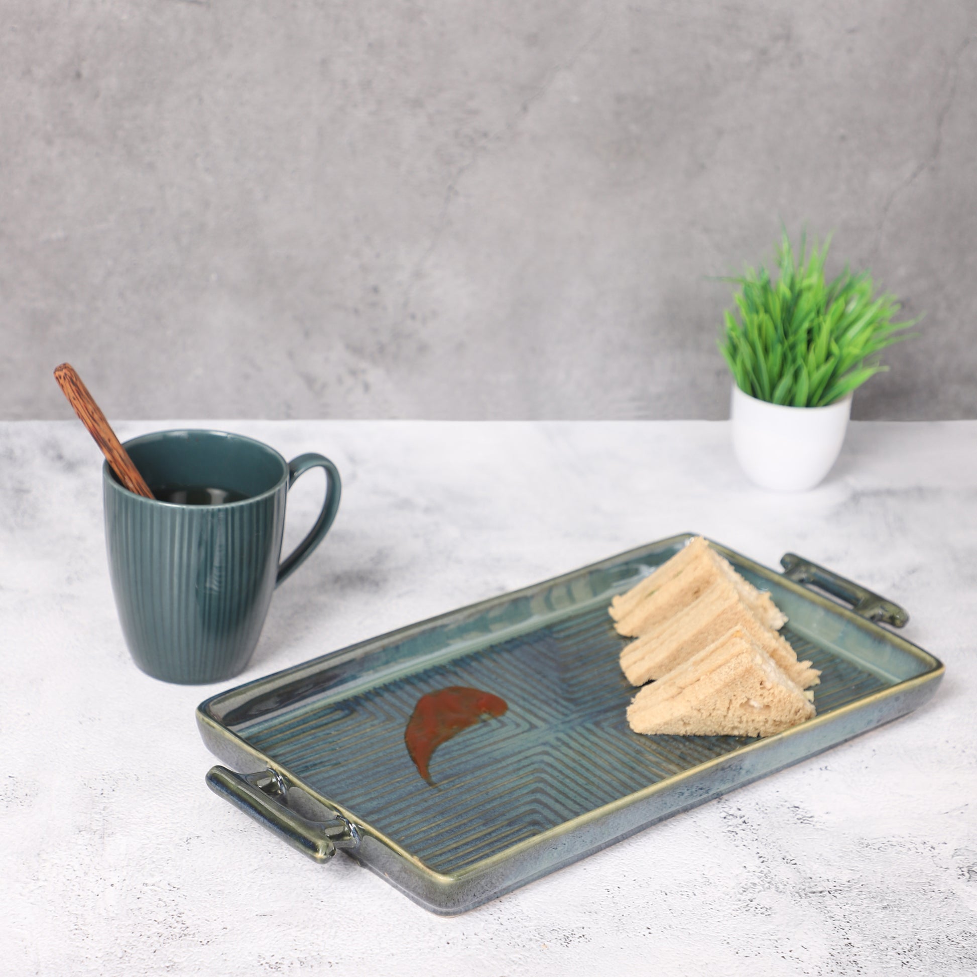 Rectangular ceramic serving tray with teal patterns, holding sandwiches, placed beside a green mug and potted plant on a table