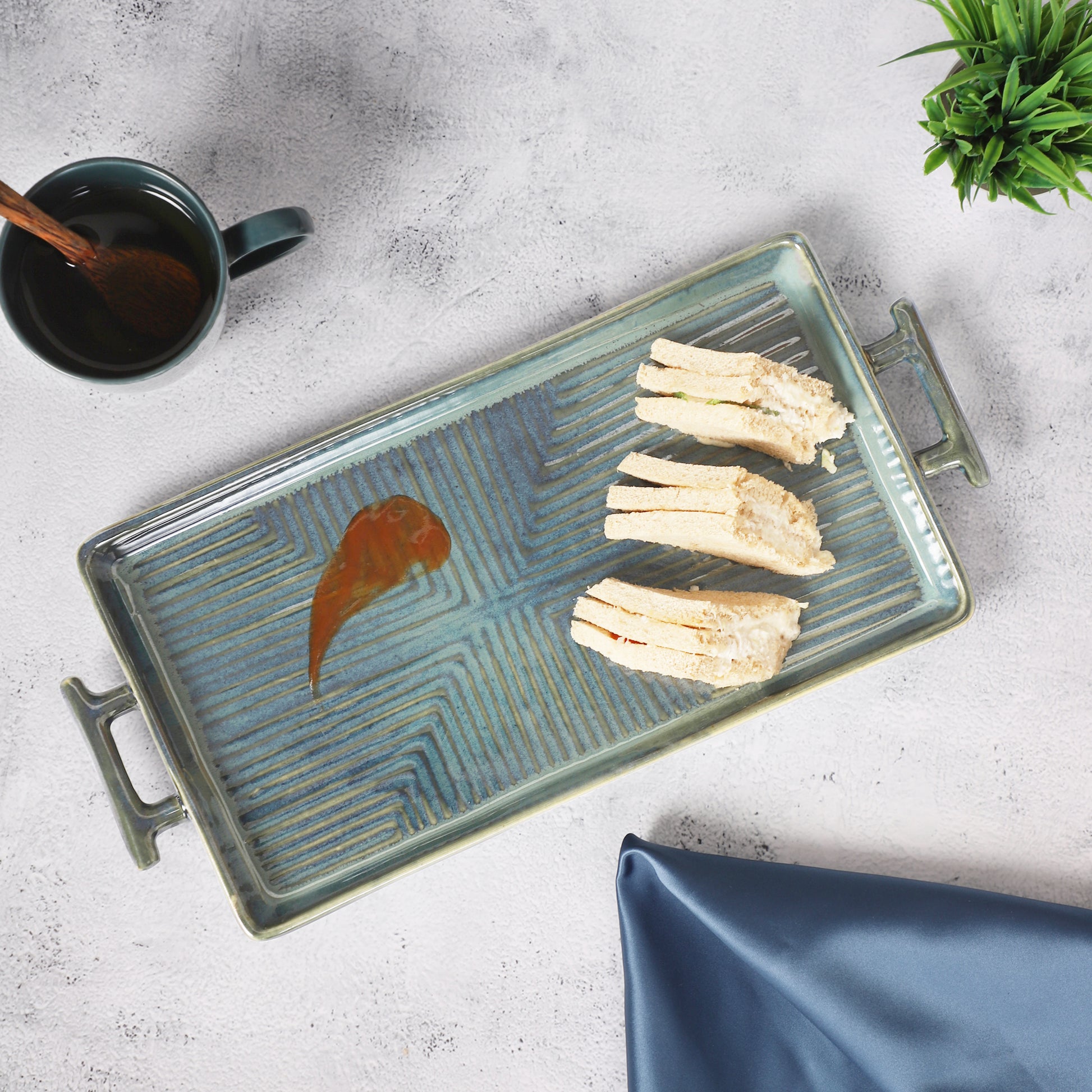 Rectangular ceramic serving tray with teal patterns, holding sandwiches, placed beside a green mug and potted plant on a table