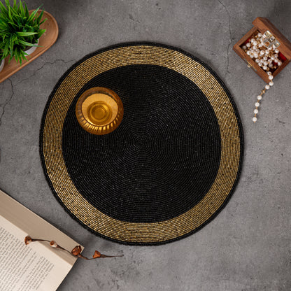Round handcrafted beaded placemat with a black center and golden border, displayed on a concrete surface alongside a gold
