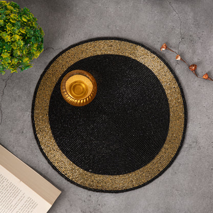 Round handcrafted beaded placemat with a black center and golden border, displayed on a concrete surface alongside a golden bowl, a vintage compass