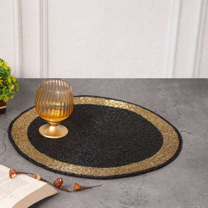 Round handcrafted beaded placemat with a black center and golden border, displayed on a concrete surface alongside a golden bowl, a vintage compass, and an open book