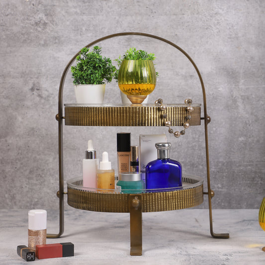 Two-tiered vintage-style metal organizer with ribbed gold finish, holding skincare bottles, a blue perfume bottle, a yellow candleholder, and small potted plants, placed on a marble surface.