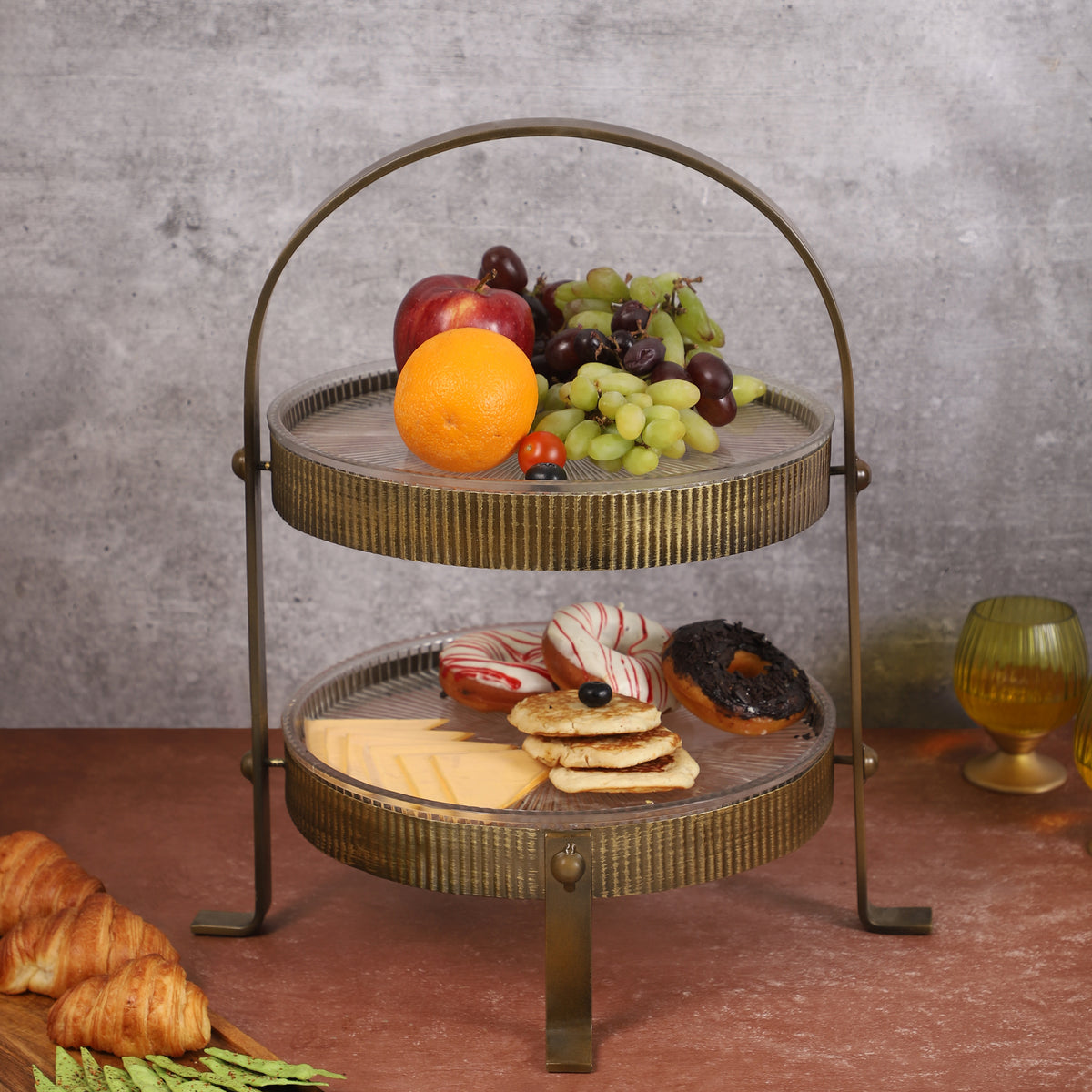 Two-tiered vintage-style metal organizer with ribbed gold finish, holding skincare bottles, a blue perfume bottle, a yellow candleholder, and small potted plants, placed on a marble surface.